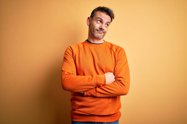 Jonge knappe man met oranje casual trui over geïsoleerde gele achtergrond lachend naar de zijkant kijkend en wegkijkend denkend. - Foto, afbeelding