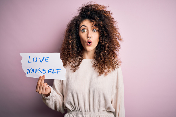 Young beautiful woman with curly hair and piercing holding paper with love yourself message scared in shock with a surprise face, afraid and excited with fear expression - Photo, Image