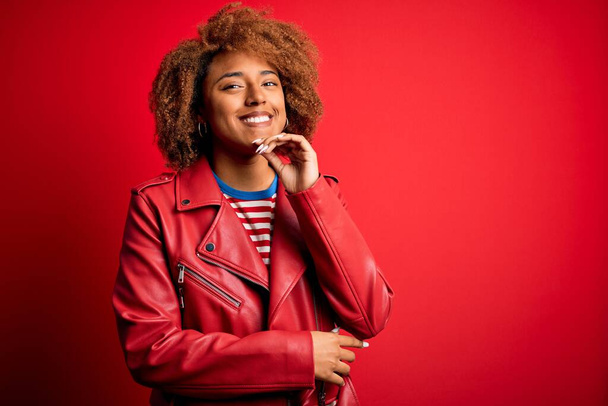 カジュアルな赤いジャケットを着た巻き毛をした若い美しいアフリカ系アメリカ人アフロ女性は、交差した腕と手をあごに上げて笑顔でカメラを自信を持って見ています。肯定的な考え. - 写真・画像