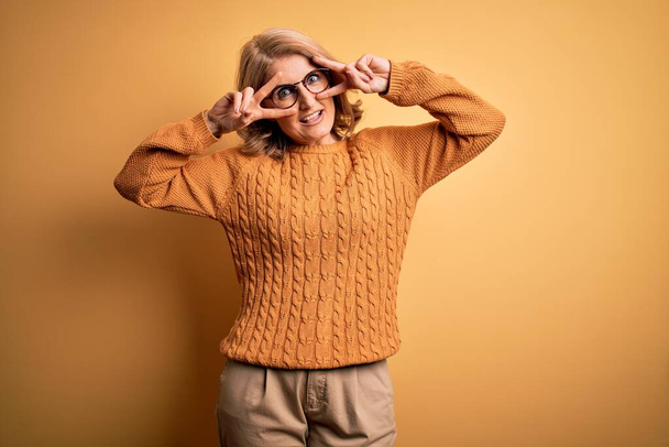 Красивая блондинка средних лет в случайном свитере и очках на желтом фоне делает символ мира с пальцами на лице, улыбаясь весело показывая победу
 - Фото, изображение