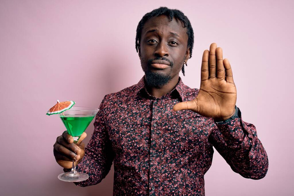 Giovane uomo afroamericano che beve cocktail verde. bevanda su sfondo rosa con mano aperta facendo segno di stop con espressione seria e sicura, gesto di difesa - Foto, immagini
