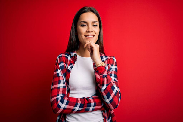 Young beautiful brunette woman wearing casual shirt standing over isolated red background looking confident at the camera with smile with crossed arms and hand raised on chin. Thinking positive. - Zdjęcie, obraz