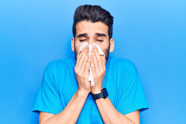 Young hispanic man illness using paper handkerchief on nose. Standing over isoltated blue background - Fotó, kép