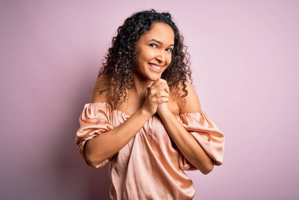 Young beautiful woman with curly hair wearing casual t-shirt standing over pink background laughing nervous and excited with hands on chin looking to the side - Фото, изображение