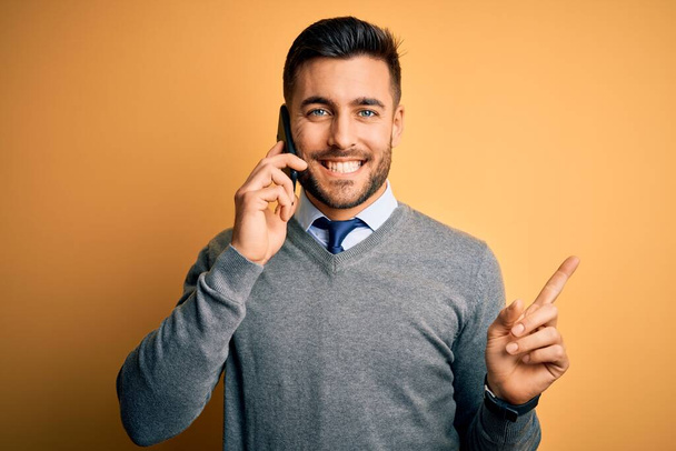Young handsome man having conversation talking on the smartphone over yellow background very happy pointing with hand and finger to the side - Foto, Bild
