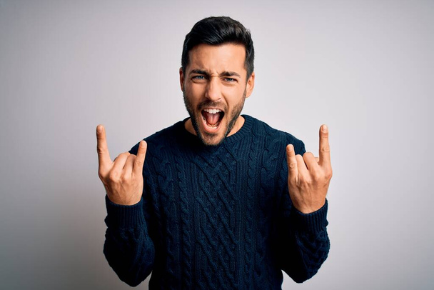 Young handsome man with beard wearing casual sweater standing over white background shouting with crazy expression doing rock symbol with hands up. Music star. Heavy concept. - Foto, Imagen