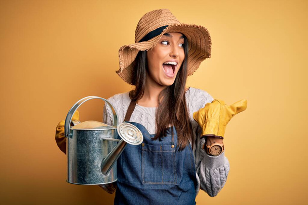 Νεαρή όμορφη μελαχρινή αγρότισσα γυναίκα φορώντας ποδιά και καπέλο κρατώντας ποτιστήρι δείχνει και δείχνει με τον αντίχειρα μέχρι το πλάι με χαρούμενο πρόσωπο χαμογελώντας - Φωτογραφία, εικόνα