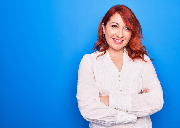 Young beautiful redhead businesswoman wearing elegant shirt standing over blue background happy face smiling with crossed arms looking at the camera. Positive person. - Fotoğraf, Görsel