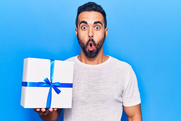 Jeune homme hispanique tenant cadeau effrayé et étonné avec la bouche ouverte pour la surprise, visage incrédule  - Photo, image