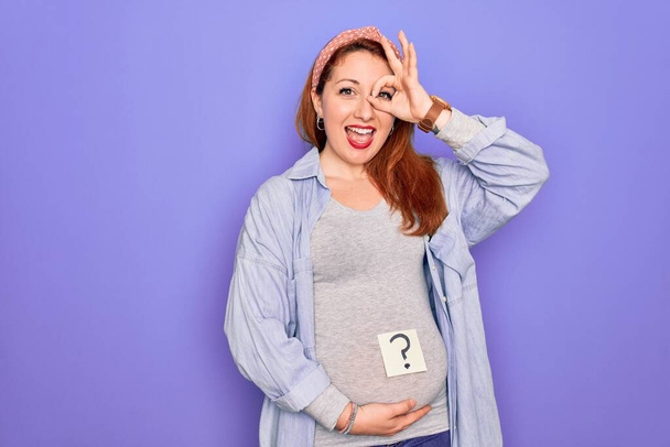 Młoda piękna ruda kobieta w ciąży spodziewa się dziecka ze znakiem zapytania na brzuchu ze szczęśliwą twarzą uśmiechnięta robi ok znak z ręki na oko patrząc przez palce - Zdjęcie, obraz