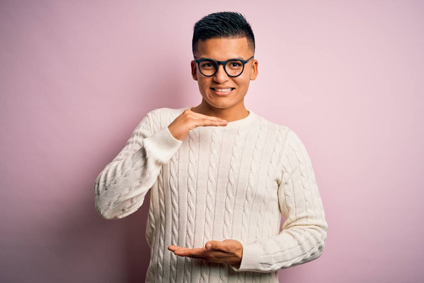 Giovane bell'uomo latino che indossa maglione casual bianco e occhiali su sfondo rosa gesticolando con le mani che mostrano segni di grandi e grandi dimensioni, simbolo di misura. Sorridendo guardando la telecamera. Concetto di misurazione
. - Foto, immagini