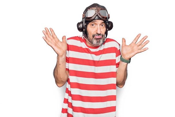 Middle age aviator man wearing vintage helmet and glasses over isolated white background clueless and confused with open arms, no idea and doubtful face. - Photo, Image