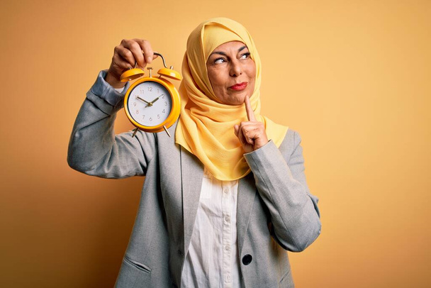 Middle age brunette business woman wearing muslim traditional hijab holding alarm clock serious face thinking about question, very confused idea - 写真・画像
