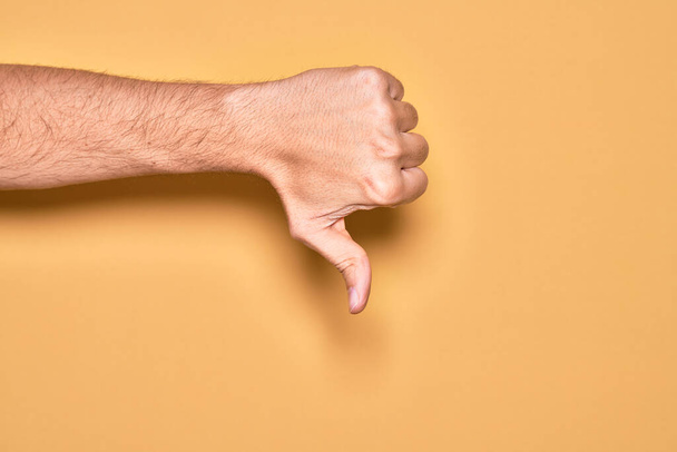 Main de jeune homme caucasien montrant les doigts sur fond jaune isolé faisant pouce vers le bas geste de rejet, désapprobation dégoût et signe négatif - Photo, image