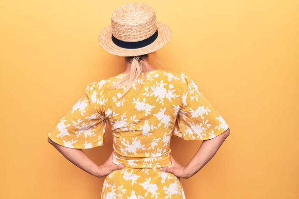 Beautiful blonde woman on vacation wearing summer hat and dress over yellow background standing backwards looking away with arms on body - Photo, Image