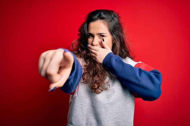 Young beautiful woman with curly hair wearing casual sweatshirt over isolated red background laughing at you, pointing finger to the camera with hand over mouth, shame expression - Foto, Imagem