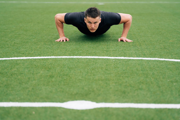 Portrait of a young male athlete in black clothes doing push-ups, look at the camera. The concept of online education of small business. Remote workout by fitness trainer - Φωτογραφία, εικόνα