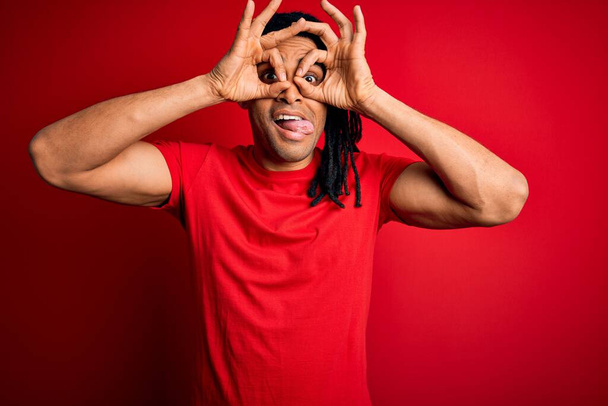Joven hombre afro afroamericano guapo con rastas que usan una camiseta casual roja haciendo un buen gesto como binoculares sacando la lengua, ojos mirando a través de los dedos. Expresión loca
. - Foto, Imagen