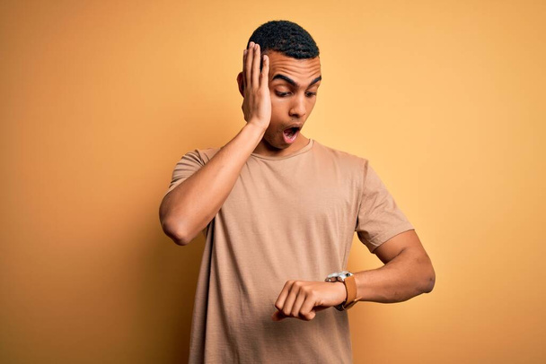 Νεαρός όμορφος Αφροαμερικάνος που φοράει casual t-shirt πάνω από κίτρινο φόντο κοιτάζοντας την ώρα του ρολογιού ανήσυχος, φοβούμενος να αργήσει - Φωτογραφία, εικόνα