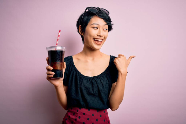 Genç, güzel Çinli bir kadın pembe arka planda saman kullanarak kola içiyor ve gülümseyen bir gülümsemeyle baş parmağıyla yüzünü gösteriyor. - Fotoğraf, Görsel