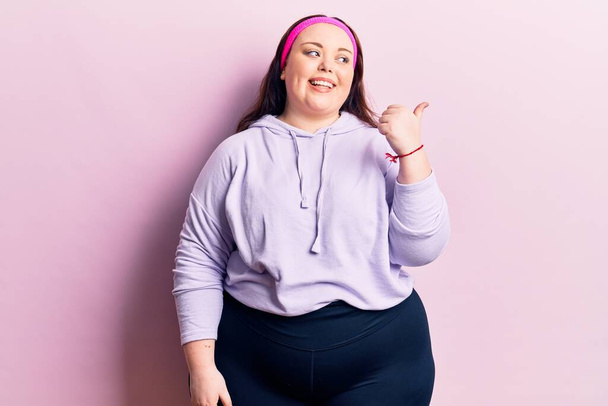 Young plus size woman wearing sportswear pointing thumb up to the side smiling happy with open mouth  - Foto, immagini