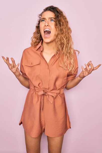 Young beautiful woman with blue eyes wearing casual summer dress over pink background crazy and mad shouting and yelling with aggressive expression and arms raised. Frustration concept. - Photo, image