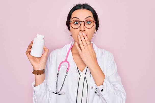 Young beautiful doctor woman with blue eyes wearing stethoscope holding medicine pills cover mouth with hand shocked with shame for mistake, expression of fear, scared in silence, secret concept - Photo, Image