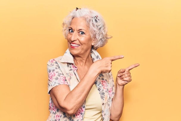 Starsza siwowłosa kobieta w luźnych ubraniach uśmiechnięta i patrząca w kamerę z dwoma rękami i palcami na boku.  - Zdjęcie, obraz