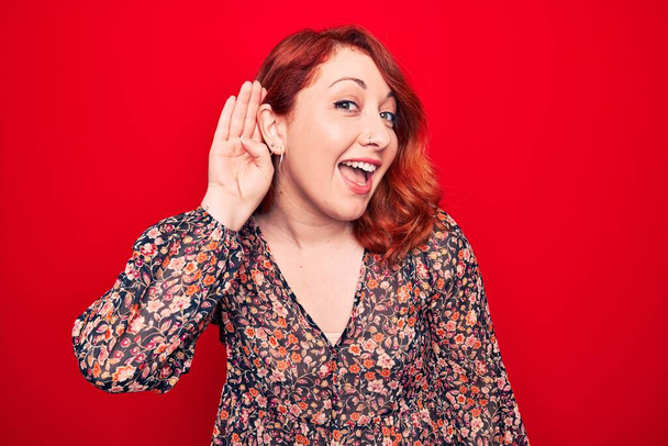 Young beautiful redhead woman wearing floral casual dress standing over red background smiling with hand over ear listening and hearing to rumor or gossip. Deafness concept. - Photo, Image
