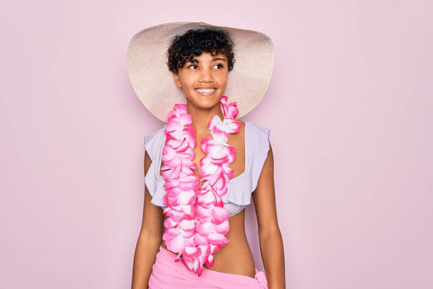 Joven hermosa mujer turista afroamericana con bikini y flores de lei hawaiano mirando hacia un lado con sonrisa en la cara, expresión natural. Riendo confiado
. - Foto, imagen