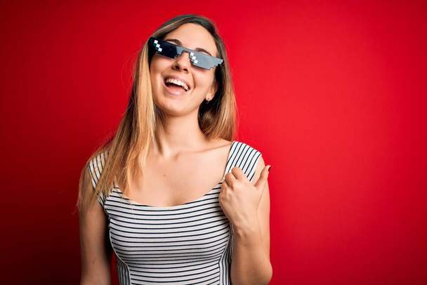 Young beautiful brunette woman wearing funny thug life sunglasses over red background smiling with happy face looking and pointing to the side with thumb up. - Zdjęcie, obraz