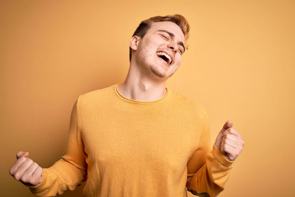 Молодой красивый рыжий мужчина в случайном свитере на изолированном желтом фоне очень счастлив и взволнован, делая жест победителя с поднятыми руками, улыбаясь и крича об успехе. Концепция празднования
. - Фото, изображение