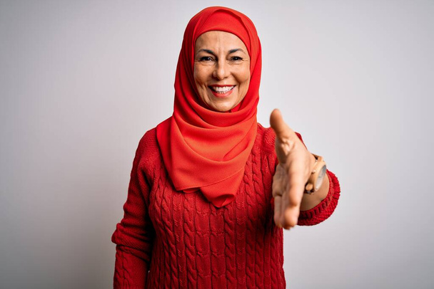 Brunetka w średnim wieku, nosząca tradycyjny hidżab muzułmański na odizolowanym białym tle, uśmiechnięta, przyjazna, oferująca uścisk dłoni jako powitanie i powitanie. Sprawy zakończone powodzeniem. - Zdjęcie, obraz
