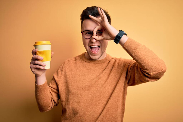 Jeune bel homme caucasien buvant du café à emporter thermo bouteille avec le visage heureux souriant faire ok signe avec la main sur les yeux regardant à travers les doigts - Photo, image
