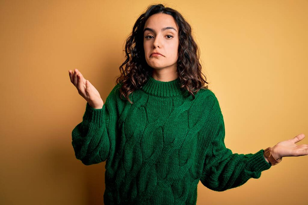 Young beautiful woman with curly hair wearing green casual sweater over yellow background clueless and confused expression with arms and hands raised. Doubt concept. - Photo, image