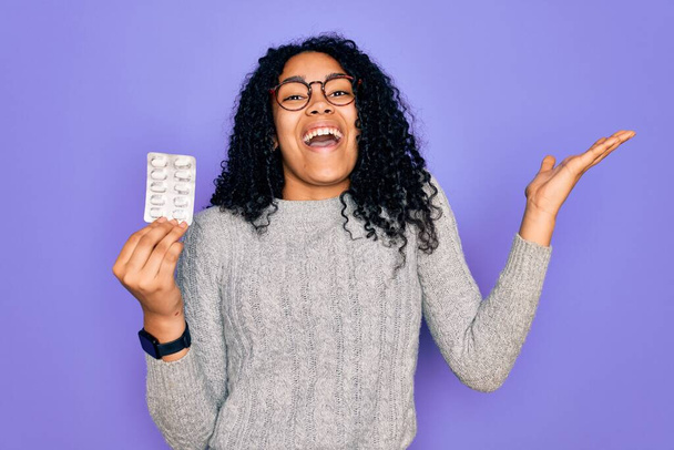 Mladý africký americký kudrnatý žena drží pilulky stojící nad izolované fialové pozadí velmi šťastný a vzrušený, vítěz výraz slaví vítězství s velkým úsměvem a zvedl ruce - Fotografie, Obrázek