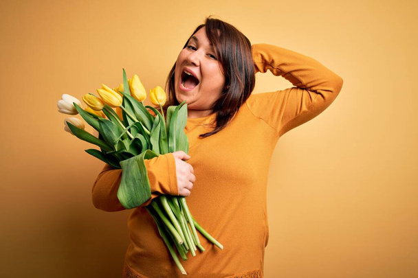 Mulher bonita plus size segurando buquê romântico de flores de tulipas naturais sobre fundo amarelo Louco e assustado com as mãos na cabeça, com medo e surpreso de choque com a boca aberta
 - Foto, Imagem