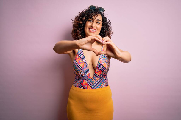 Joven mujer árabe hermosa de vacaciones con traje de baño y gafas de sol sobre fondo rosa sonriendo en el amor haciendo forma de símbolo del corazón con las manos. Concepto romántico
. - Foto, imagen