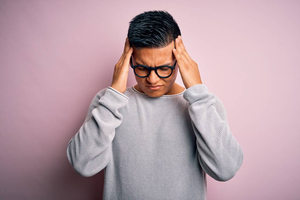 Hombre latino guapo joven que usa suéter casual y gafas sobre fondo rosa que sufre de dolor de cabeza desesperado y estresado porque el dolor y la migraña. Manos en la cabeza
. - Foto, imagen