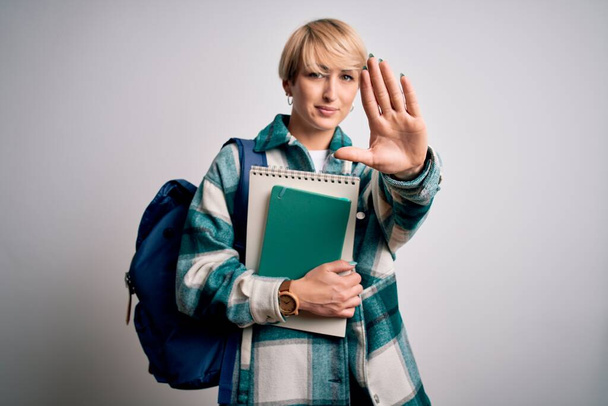 Young blonde student woman with short hair wearing backpack and holding university books with open hand doing stop sign with serious and confident expression, defense gesture - Фото, изображение