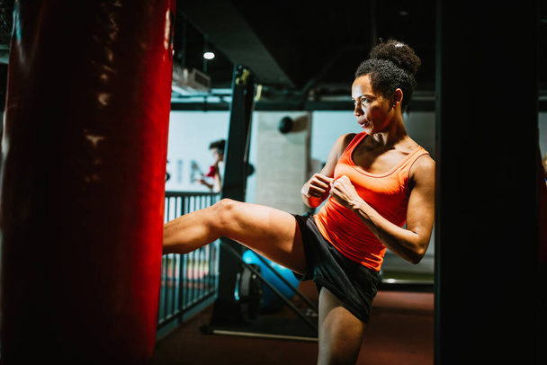Beautiful black fit woman in sportswear boxing kicking bag in the gym. - Photo, Image