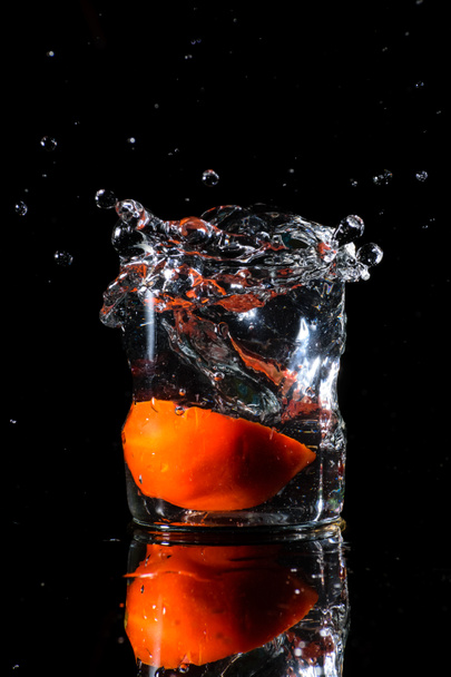 A glass of water in a dark background and water being splashed with a piece of cut tomato dropped inside the glass - Foto, immagini