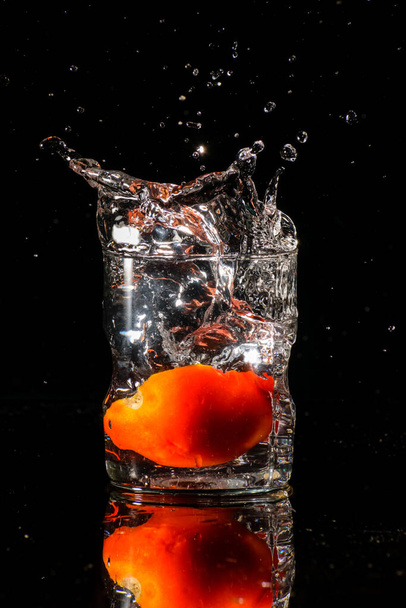暗い背景に水のガラスと水がガラスの中に落ちるカットトマトの一片で発射されています - 写真・画像