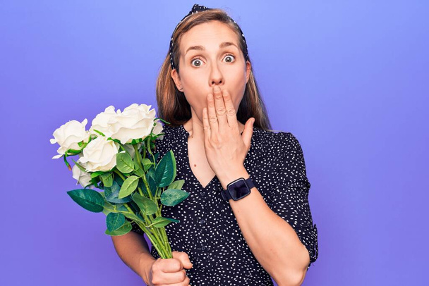 Young beautiful blonde woman holding romantic bouquet of white roses covering mouth with hand, shocked and afraid for mistake. surprised expression  - Photo, Image