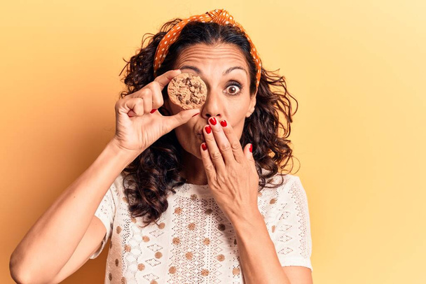 Orta yaşlı, güzel bir kadın, çikolatalı kurabiyeyi gözünün üstüne tutarak ağzını eliyle kapatıyor, şok geçiriyor ve hata yapmaktan korkuyor. şaşırmış ifade  - Fotoğraf, Görsel