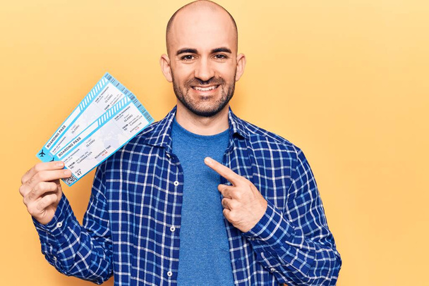 Young handsome bald man holding airline boarding pass smiling happy pointing with hand and finger  - Photo, Image