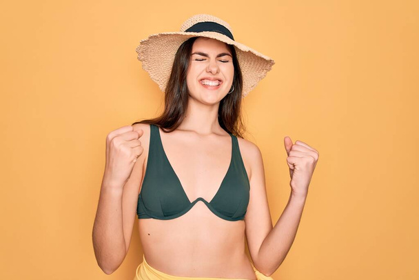 Young beautiful girl wearing swimwear bikini and summer sun hat over yellow background excited for success with arms raised and eyes closed celebrating victory smiling. Winner concept. - Photo, Image