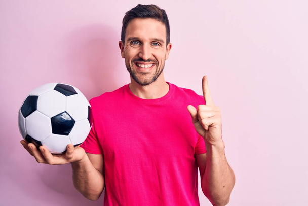Jovem jogador bonito homem jogando futebol segurando bola de futebol sobre fundo rosa isolado sorrindo com uma ideia ou pergunta apontando dedo com cara feliz, número um
 - Foto, Imagem