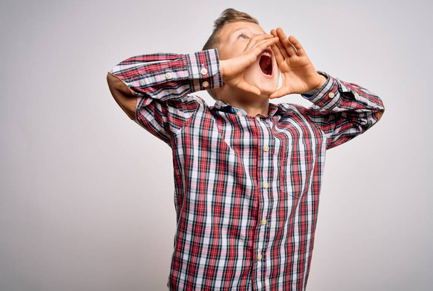 Νεαρό μικρό καυκάσιο παιδί με μπλε μάτια φορώντας κομψό πουκάμισο στέκεται πάνω από απομονωμένο φόντο Φωνάζοντας θυμωμένος δυνατά με τα χέρια πάνω από το στόμα - Φωτογραφία, εικόνα