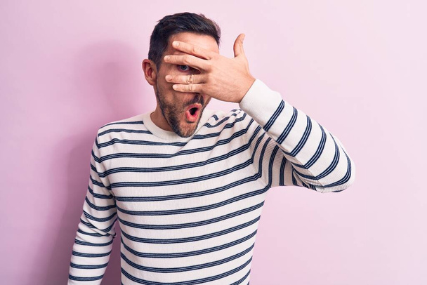 Mladý pohledný muž v neformálním pruhovaném svetru, stojící nad izolovaným růžovým pozadím, vykukující v šoku a zakrývající obličej a oči rukou, hledící skrz prsty ustrašené - Fotografie, Obrázek